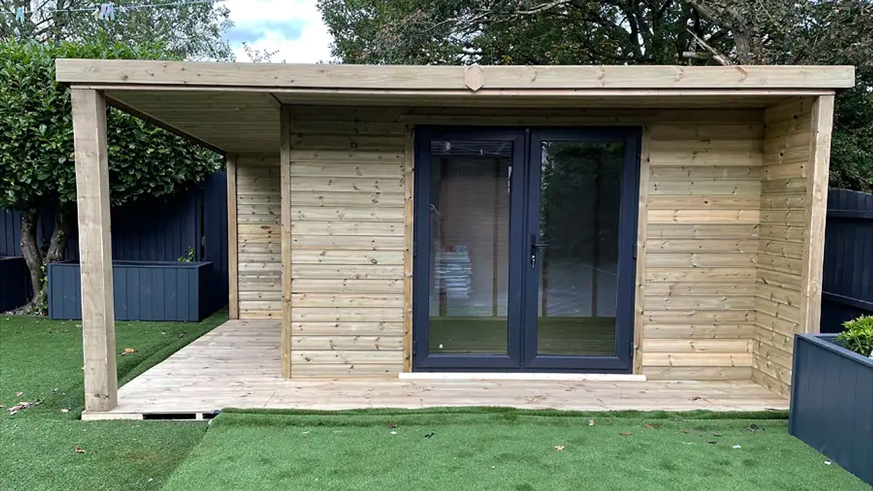 A pent roof timber garden room with glazed double doors and a covered decked seating area on a neat lawn