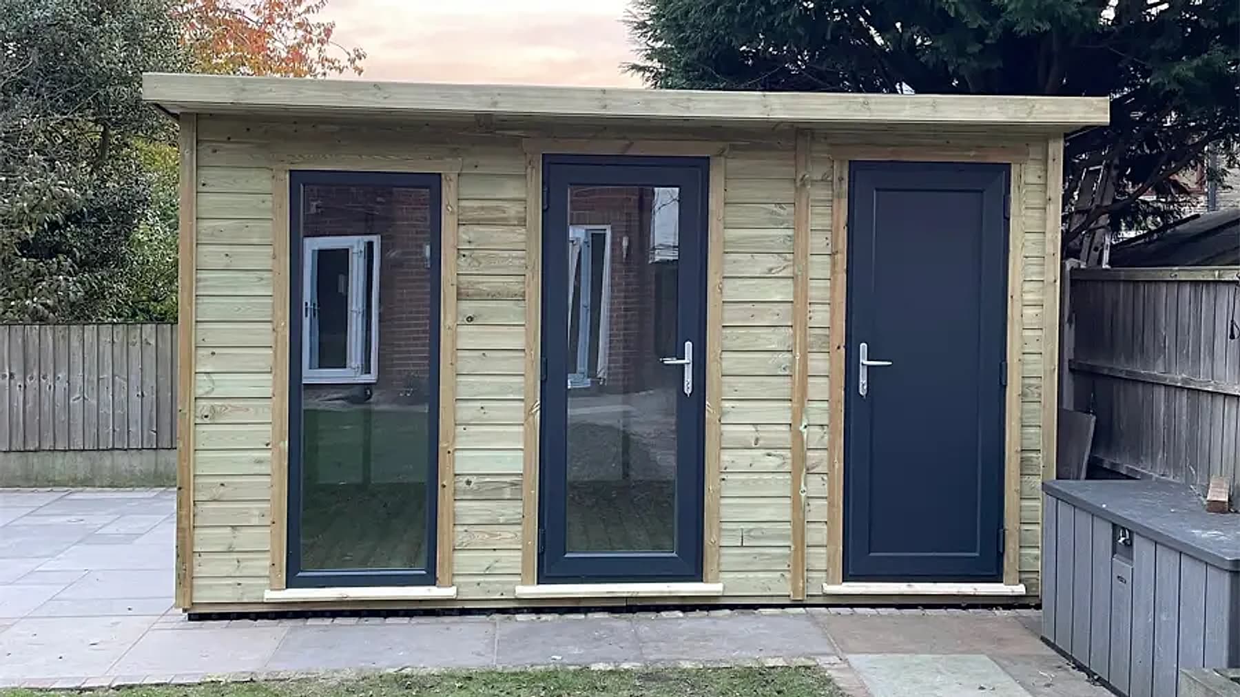A pent roof timber summer house and shed combination with a window and two doors on a paved patio