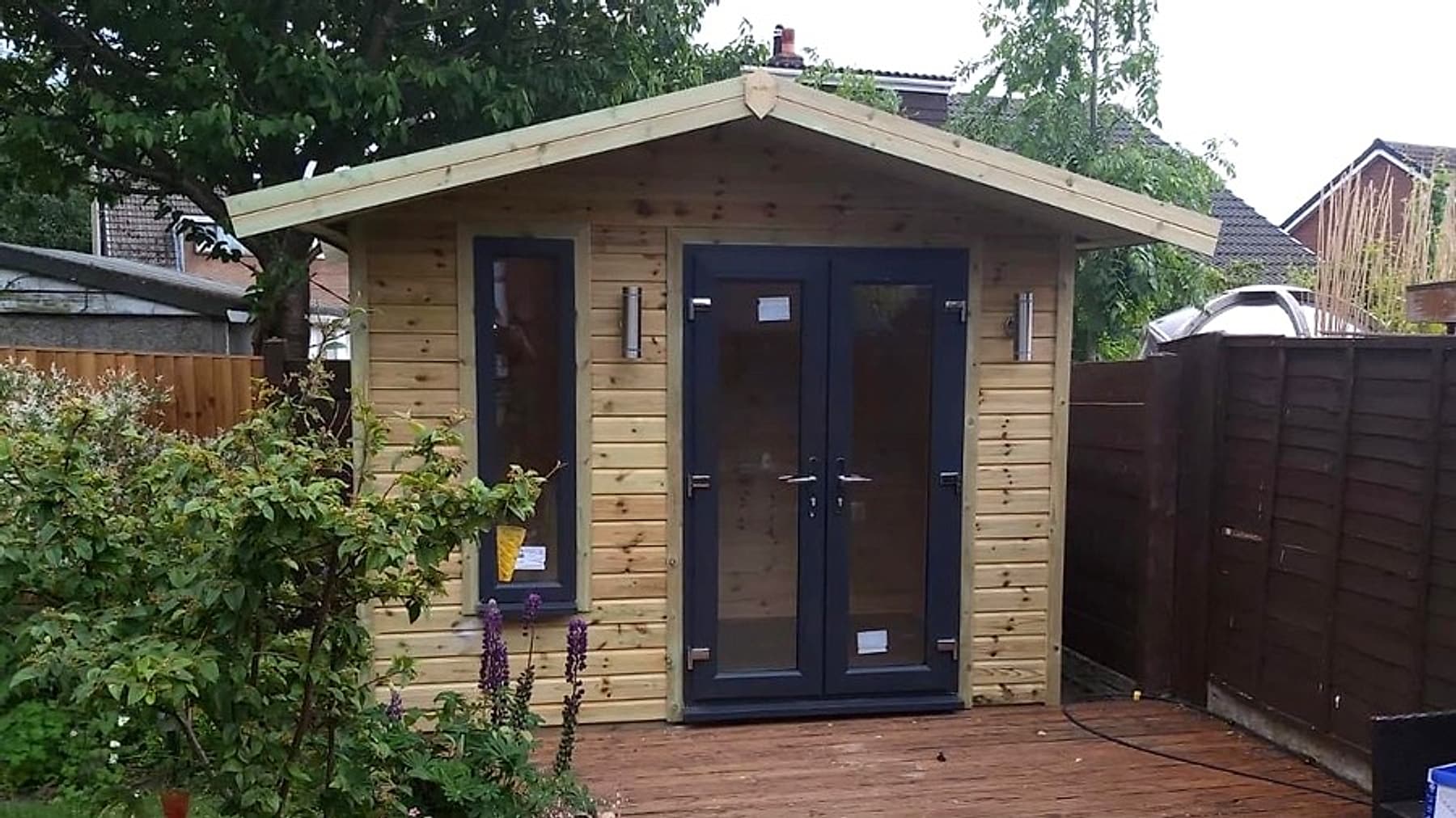 A gabled timber chalet-style summer house with grey double doors, set on decking with planting around it