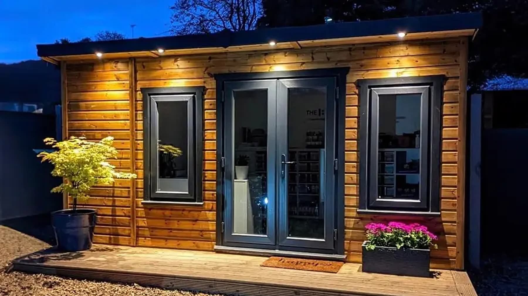 A timber summer house at dusk, lit by spotlights above grey double doors, with a golden acer in a pot and pink flowers